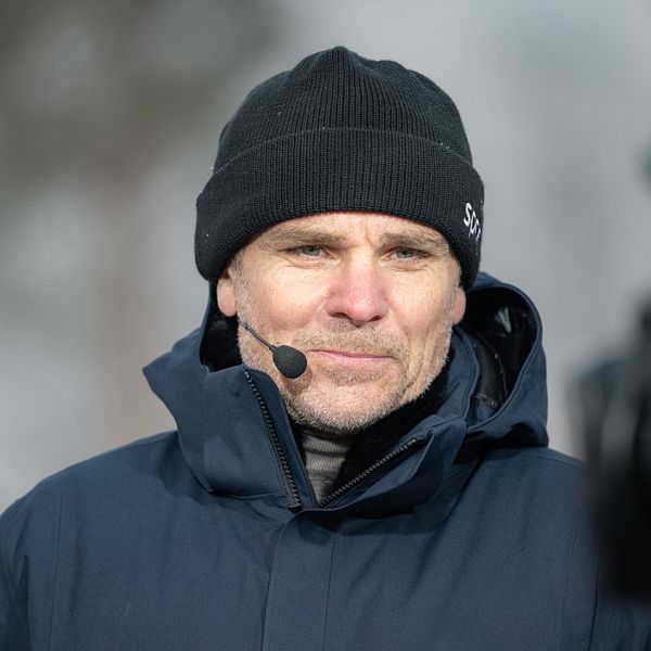 Person i mörk vinterjacka och mössa utomhus, träd syns suddigt i bakgrunden.