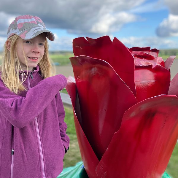 Flicka i tioårsåldern i lila jacka står bredvid en stor röd rosstaty utomhus.