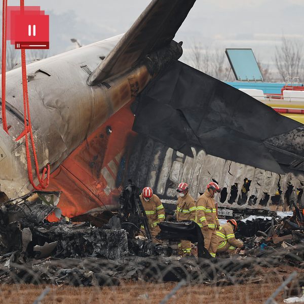Brandmän inspekterar vraket av ett kraschade flygplan som har tagit skador av brand, med omsorgsfullt arbete i närheten av det förstörda fordonet. En rad räddningstjänstfordon syns i bakgrunden.