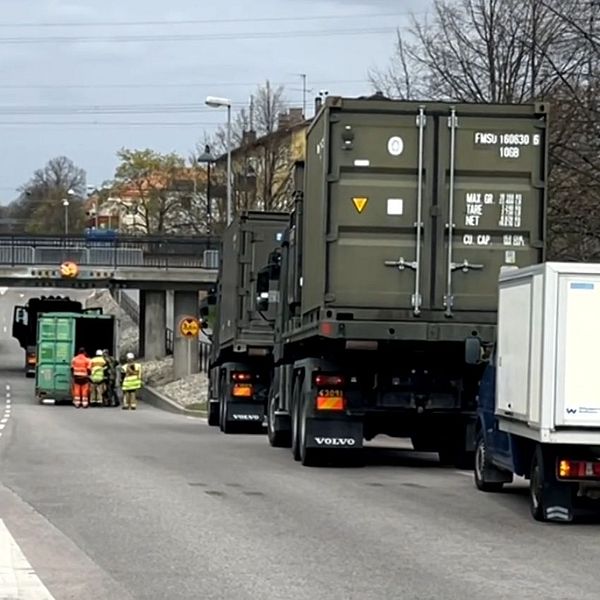 Flera lastbilar, inklusive militärfordon, blockerar en väg under en bro, arbetare på plats.