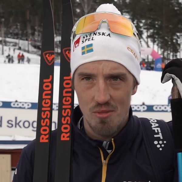 En längdskidåkare iförd mössa och jacka med SVT-loggor håller i ett par skidstavar. I bakgrunden syns en snötäckt skidarena med en annan skidåkare och träd.