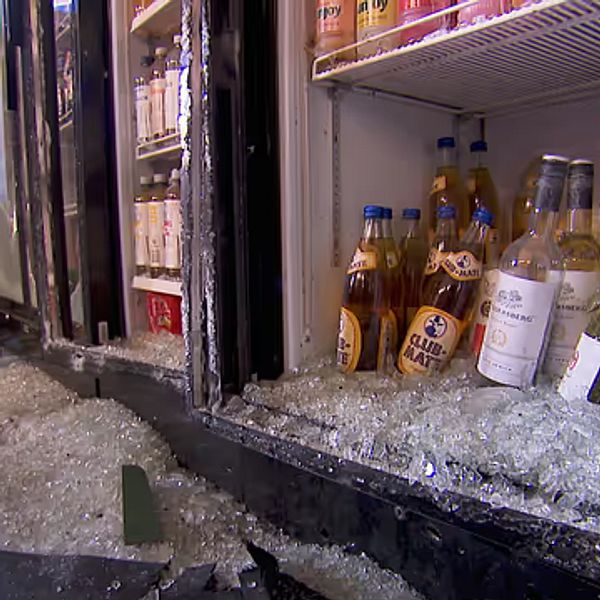 Flaskor bland krossat glas framför trasigt kylskåp i butik.