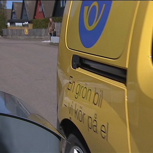 En elbil i gult står parkerad vid en väg. I bakgrunden syns en asfaltgata med hus längs sidan. På bilen finns texten ”En grön bil – vi kör på el”. En grå postlåda syns i förgrunden.