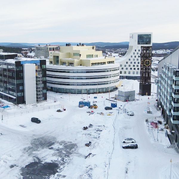 Vy över en snöklädd byggarbetsplats med flera moderna byggnader i en stadsmiljö. Det är delar av Kirunas nya centrum. I mitten syns en stor, rundad byggnad med en ljus färg, medan omgivande byggnader har en mer traditionell arkitektur. Snön täcker marken och det finns byggmaskiner samt annat arbetsmaterial synligt. I bakgrunden kan man se fler stadsbyggnader och en vidsträckt himmel.