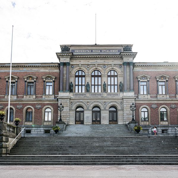 Universitetshuset i Uppsala, med en stor trappa som leder upp till byggnadens monumentala fasad, omgiven av flaggstänger och blomkrukor. Två personer går på trappan.