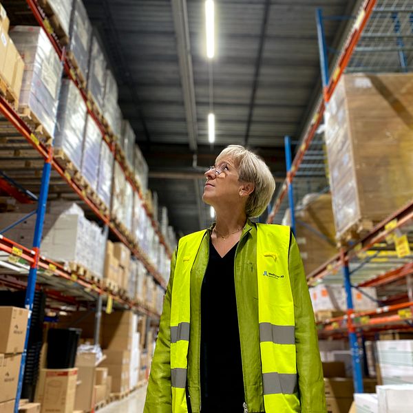 Person i gul väst står i ett lager mellan höga hyllor fyllda med kartonger och paket.