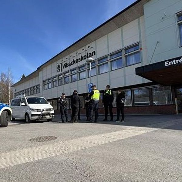 Polis och civilfordon står utanför entré till en byggnad under blå himmel.