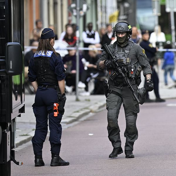 Poliser står på en gata där en incident har inträffat. En beväpnad polis i taktisk utrustning går framåt, medan en annan polis övervakar situationen. I bakgrunden syns en folkmassa och ett polisfordon. Området är avspärrat med avspärrningsband.