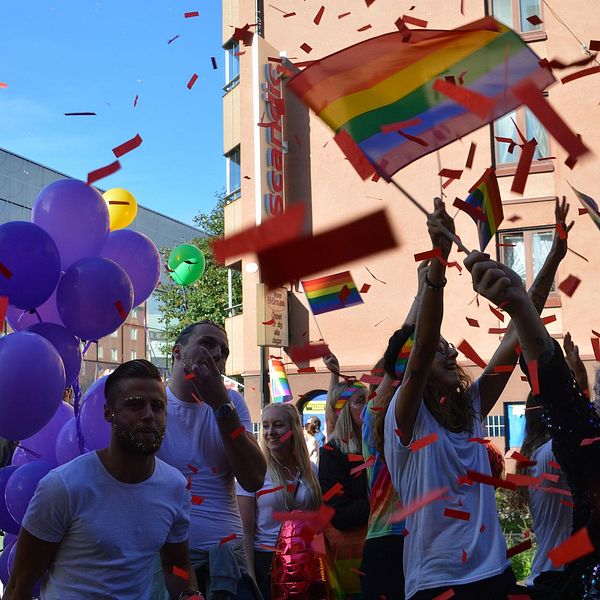 Personer firar Pride med regnbågsflaggor och lila ballonger. Confetti i luften.