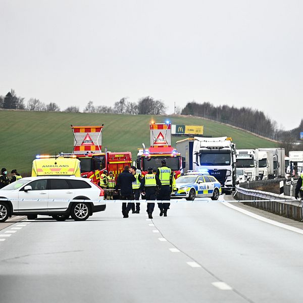 En motorväg som blockerats av räddningstjänst och polis efter endöd man påträffats i höjd med Skrea. Flera fordon, inklusive lastbilar och ambulans, syns i bakgrunden. Räddningsteam arbetar på platsen, medan poliser dirigerar trafiken. Målet med insatsen tycks vara att hantera olyckan och säkerställa trafiksäkerheten.