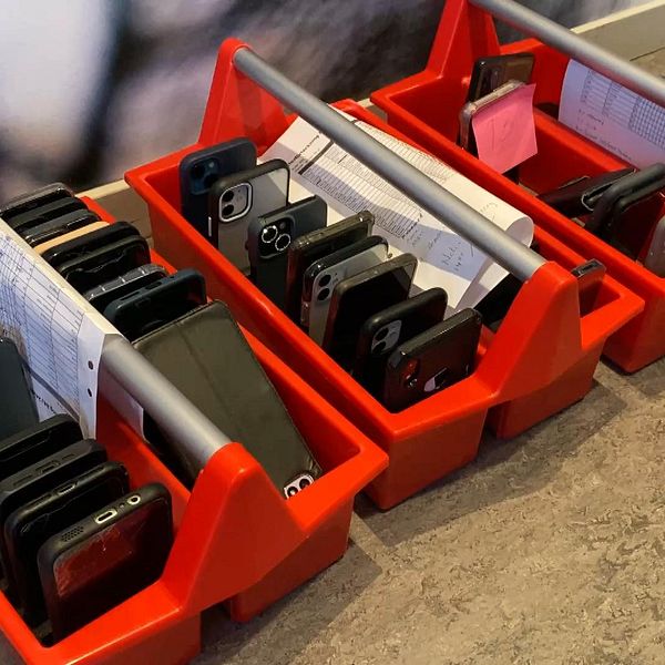 Två röda korgar med flera mobiltelefoner inlagda, ordnade i rader. Den vänstra korgen innehåller främst svarta och grå telefoner medan den högra korgen har telefoner i olika färger. Båda korgarna har också papper som delvis syns ovanpå telefonerna.
