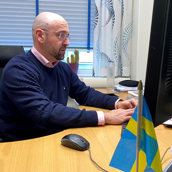 Timur Uman, professor i företagsekonomi vid högskolan i Jönköping, sitter vid ett skrivbord i ett kontorslandskap, klädd i en blå tröja. Han använder en dator med en stor skärm. På skrivbordet finns en mus och ett par små svenska flaggor. I förgrunden syns även en dekoration med traditionell svensk klädsel. Rummet är upplyst av naturligt ljus som kommer från fönstren bakom honom.