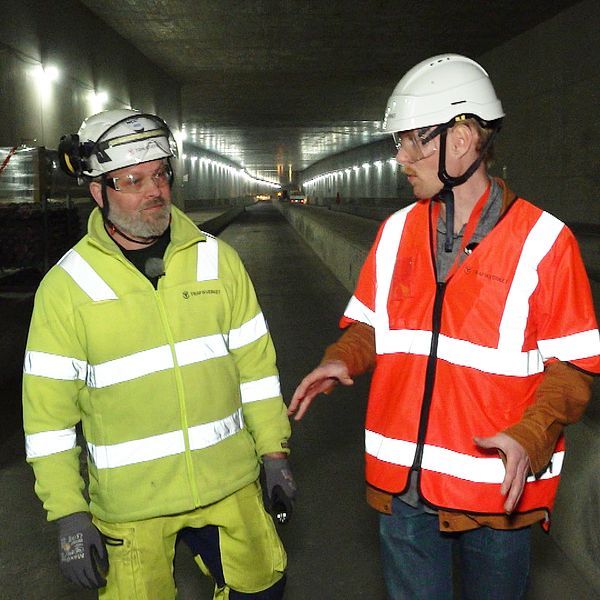 Jimy Andersson, byggledare för Västlänken vid centralstationen i Göteborg, till vänster i en grön reflexjacka. Carl Malmborg, reporter för SVT Nyheter Väst, till höger i orange reflexväst. De båda går och pratar i en lång tunnel som är en del av Västlänken.