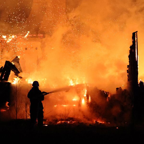 En brandman sprutande vatten på en intensiv brand som har gripit tag i en byggnad. Flammor och rök fyller luften, och omgivningen är upplyst av den starka elden. I bakgrunden syns förvridna och brinnande trästrukturer.