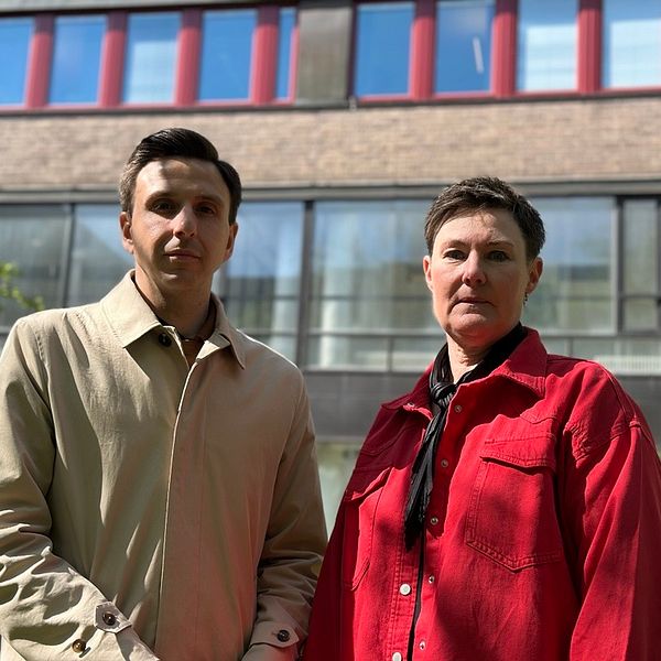 Två personer står framför en byggnad med stora fönster och rödbruna fönsterkarmar.