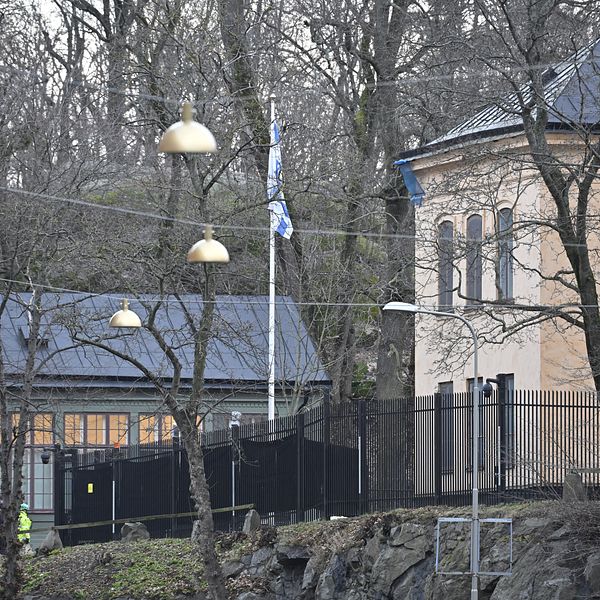 Byggnad med flaggstång omgiven av träd, en person i reflexkläder syns i förgrunden.