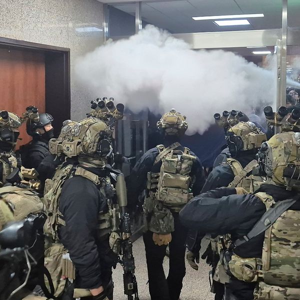 En grupp maskerade och beväpnade specialförband rör sig genom en rökfylld korridor, med dämpade färger och fokuserade uttryck. Rök strömmar från ett rum till vänster och skapar en intensiv och dramatisk atmosfär. Bakgrunderna visar stängda dörrar och en skylt för handikapptoalett i närheten.