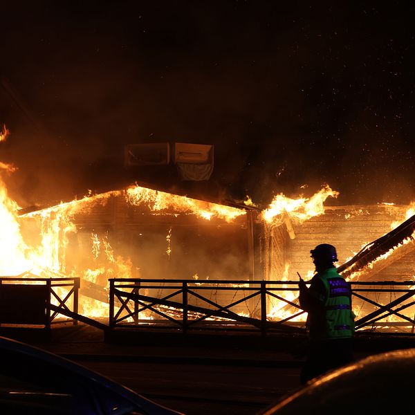 En byggnad står i lågor på natten, en brandman syns i förgrunden.