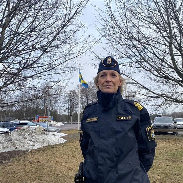 En polis i uniform står på en gräsyta med parkerade bilar i bakgrunden och en svensk flagga som vajar. Träd utan löv omger platsen, vilket antyder att det är vinter eller tidig vår. Snö kan ses samlad på marken.