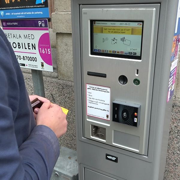 En person står vid en automat för betalning av parkering, där de håller en biljett eller kort i handen. Automaten har en digital skärm som visar olika options för betalning och instruktioner. I bakgrunden syns en informationsskylt om parkering.