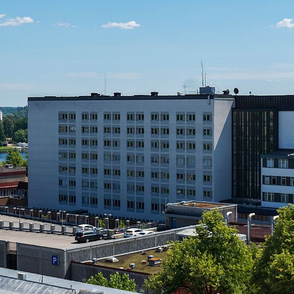 Sjukhusbyggnad omgiven av träd och en sjö i bakgrunden under en klarblå himmel.