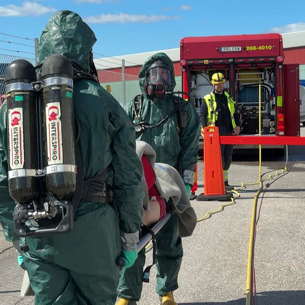 Räddningspersonal i skyddsdräkter bär en person under en övning vid industrilokal.