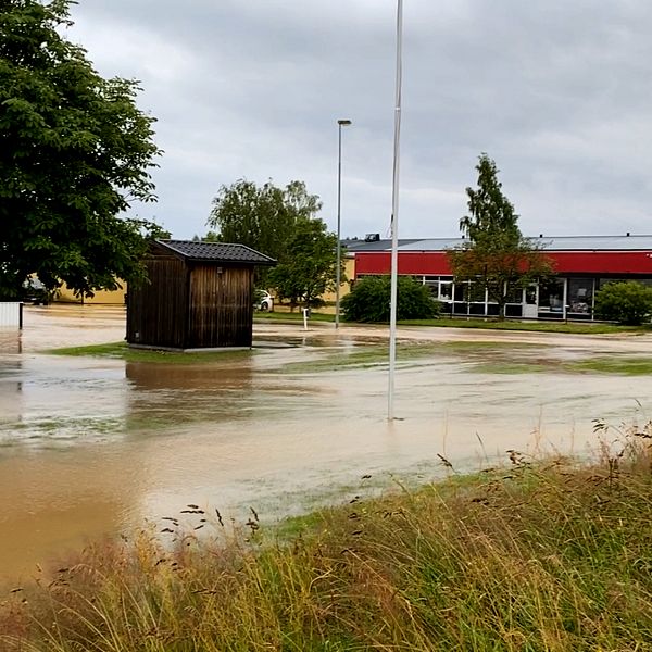 Översvämmad gata vid industribyggnad, gräsfält och träd under molnig himmel.