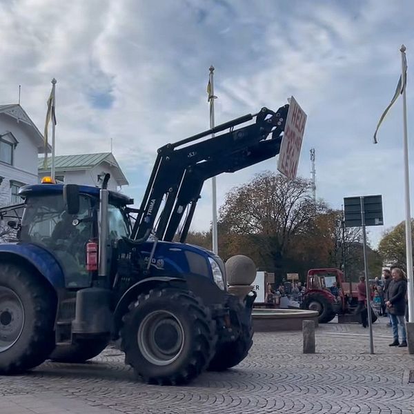 En blå traktor med en lyftanordning framför en folkmassa som samlats på en torg. Bakgrunden visar både moderna och äldre byggnader samt flera flaggstänger med flaggor. Traktorn bär ett skylt som inte är tydligt synlig, och det är en klar himmel med några moln.