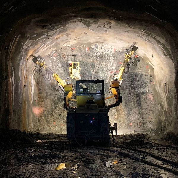 En gruvmaskin står i en mörk tunnel med ljust upplysta väggar, där den används för att bryta mineraler. Maskinens armar sträcker sig framåt och skapar ett fokus på dess arbete i den avlägsna miljön.