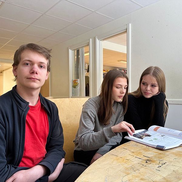 Tre personer sitter runt ett bord och tittar i en skolbok. En ljus korridor sträcker sig i bakgrunden.
