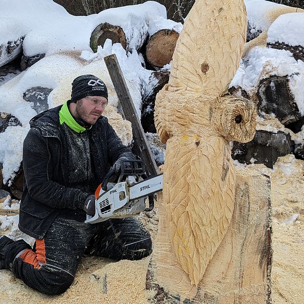 Motorsågsskulptören Johan Karlsson sitter på knä med en motorsåg i händerna och formar en stor trästock till en uggla