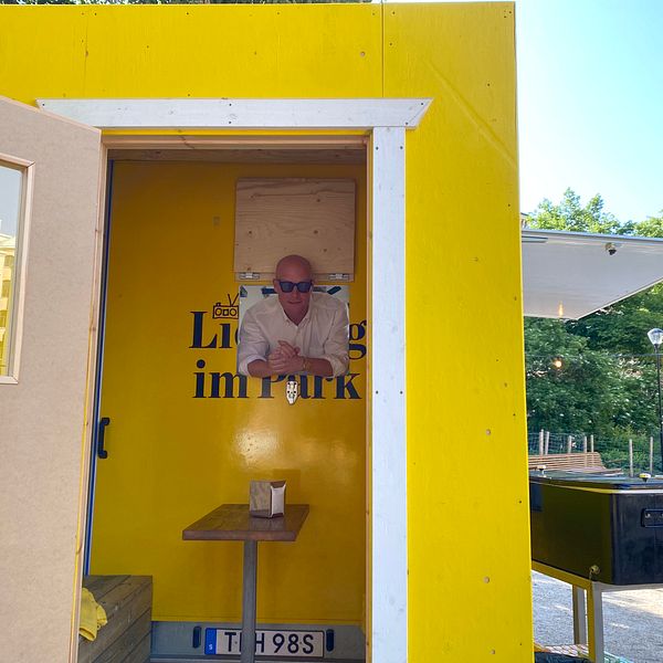 Man i solglasögon står i dörröppningen till ett litet gult hus på en solig dag.