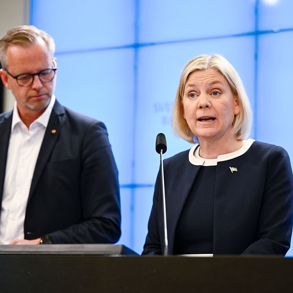 Två personer står vid ett podium under en presskonferens med en digital skärm i bakgrunden.