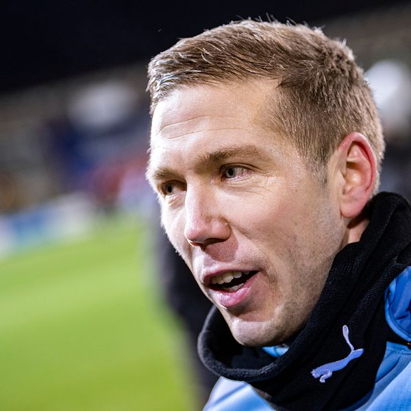 Person i ljusblå tröja står på en fotbollsplan med suddig publik i bakgrunden.