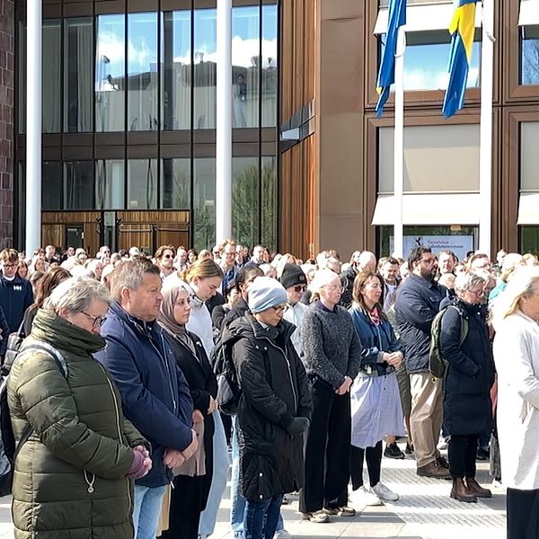 Stilla folkmassa står samlade utanför byggnad med svenska flaggor under en tyst minut.