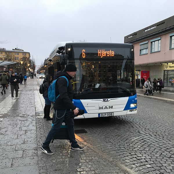 En kollektivtrafikbuss, nummer 6 mot Hjärsta, står parkerad i mitten av bilden i stadsmiljö, framför bussen passerar en person.