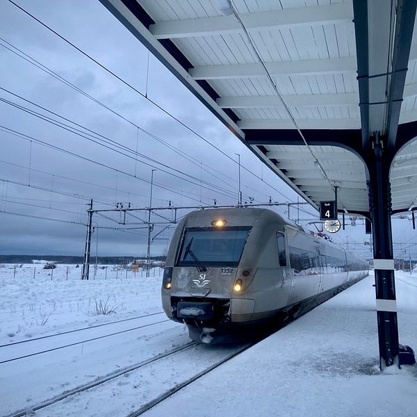 Ett modernt tåg anländer till en snöig perrong med en ensam passagerare stående bredvid spår 3. Himlen är mulen och omgivningen täcks av ett vinterlandskap.
