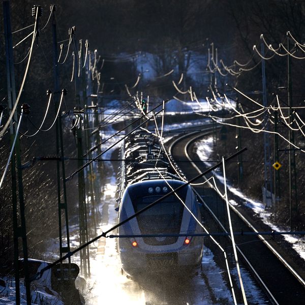 Ett persontåg åker längs en kurviga järnvägslinje i ett vintrigt landskap, omgiven av snötäckta träd och elektriska stolpar. Dämpad belysning skapar en mystisk atmosfär.