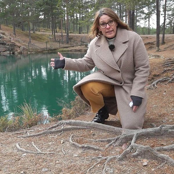 Kvinna i beige kappa och brandgula byxor står på knä framför en grön sjö i en skog