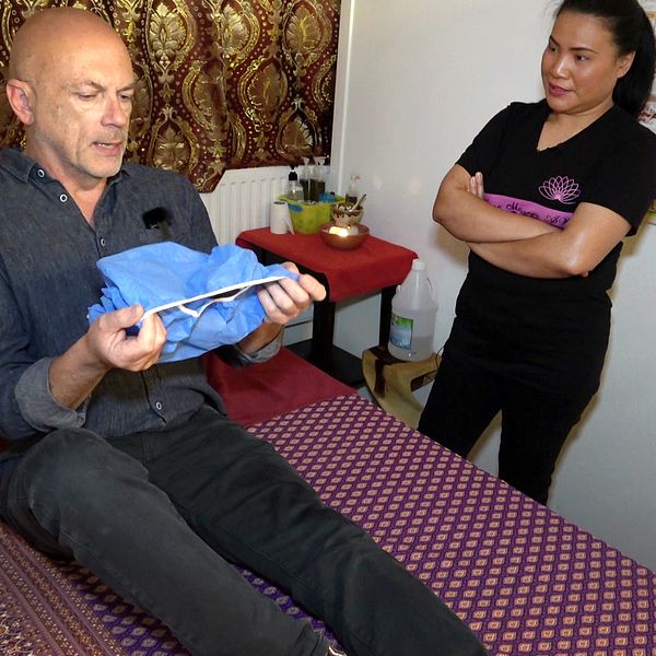 En man sitter på en massagebänk och håller en blå handduk i sina händer. I bakgrunden står en kvinna med armarna i kors. Rummet är inrett med en mönstrad kudde och en dekoration på ett bord, samt en vägg prydd med bilder.