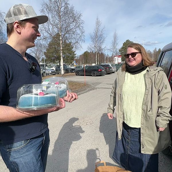 En kvinna står vid en bil och en man står och håller i två tårtor. Personerna ler mot varandra.