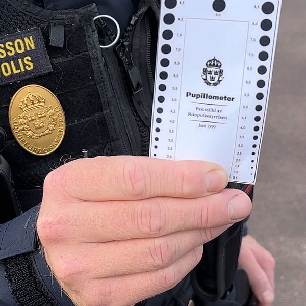 En trafikpolis håller upp en pupillometer, ett verktyg för att mäta pupilens storlek, med text som visar att det används av Rikspolisstyrelsen. Polisen är klädd i uniform med en namnskylt som tydligt anger att personen är P. Magnusson.