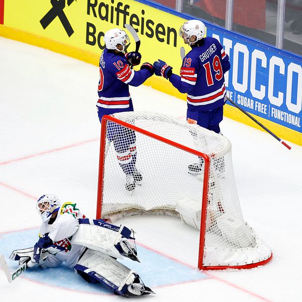 Två hockeyspelare firar mål bakom målbur, målvakt sitter på isen framför dem.