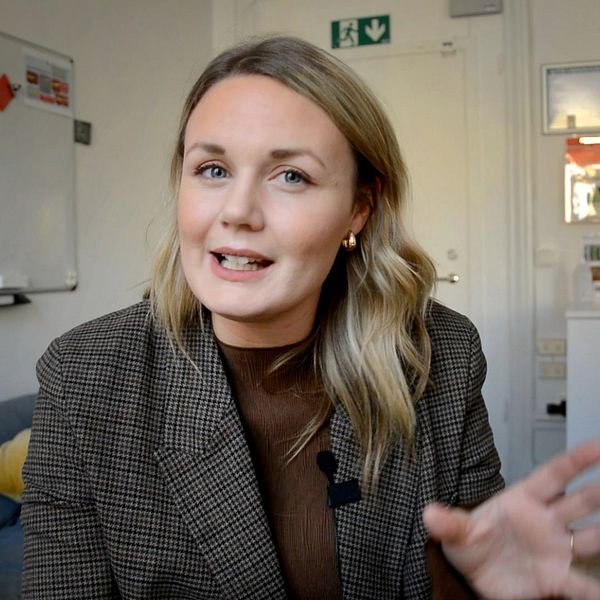 En person sitter i en modern inomhusmiljö, iförd en rutigt kavaj, och gestikulerar med en hand. I övre vänstra hörnet finns en grafik med siffrorna 3, 2, 1 i rött.