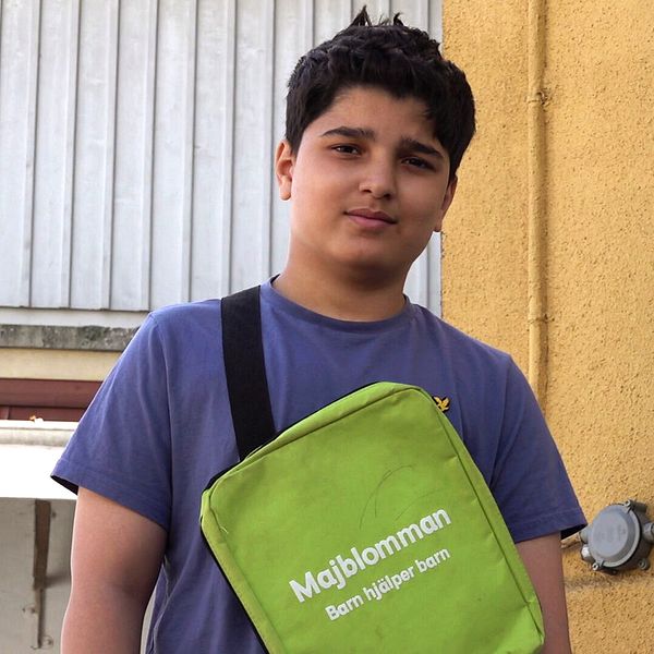 Person med grön väska märkt ”Mobilkameran” står mot en husvägg utomhus.