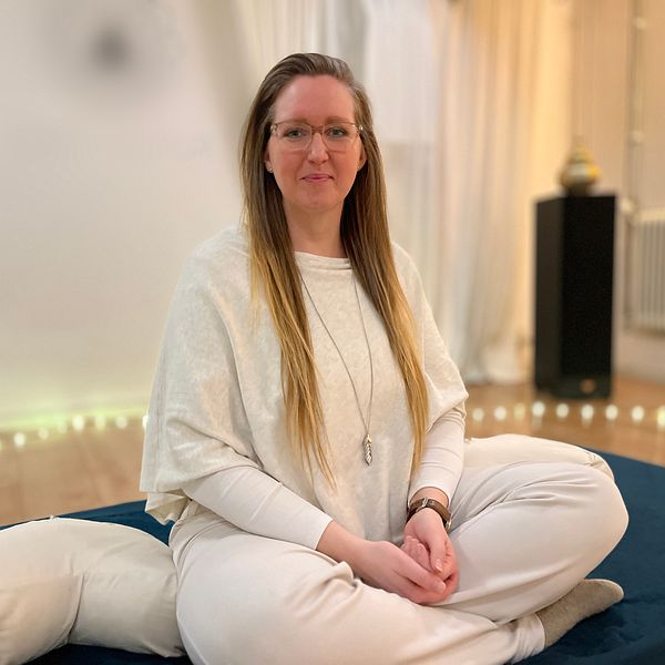 En person sitter i en meditationsställning på en blå matta i ett rum med dämpad belysning. Personen har på sig vita kläder och en lång halskedja. Bakgrunden visar ljusa gardiner, en upplyst växt och ett element.