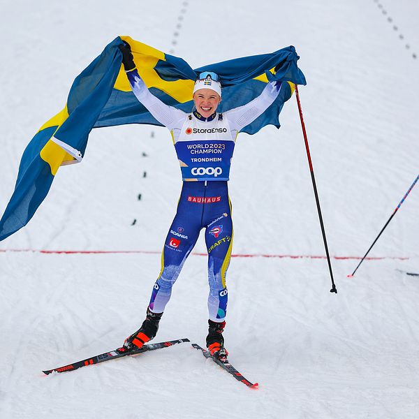 Svensk längdskidåkare firar med flagga, vid mållinjen under tävling.