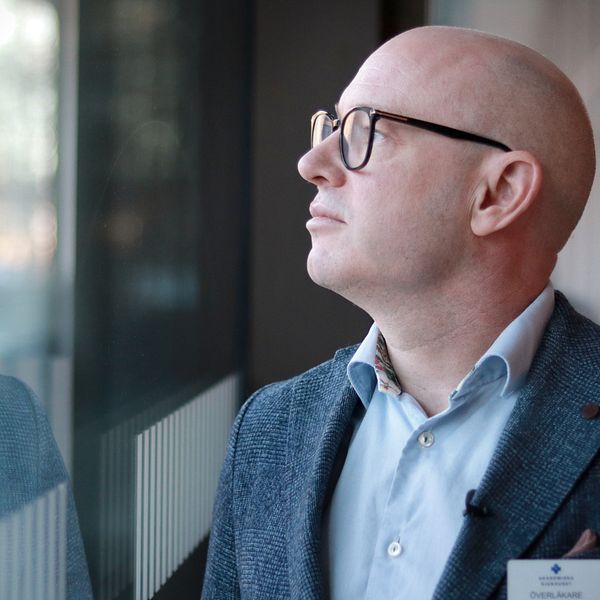 En person i en grå blazer står vid ett fönster och ser ut mot utsidan, med reflektioner synliga på glaset. Personen har en namnskylt fäst på blazern. Bakgrunden visar suddiga former av det som kan vara en modern miljö.