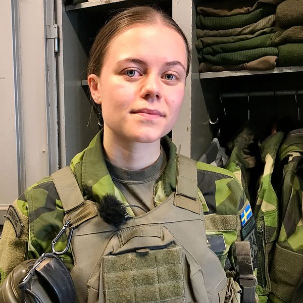 En person i militäruniform står i en klädkammare med grön och svart kamouflage. I bakgrunden syns flera militärplagg som hänger prydligt. Personen bär en mikrofon och har en Sveriges flagga på sin uniform, vilket indikerar att de tillhör den svenska militären.
