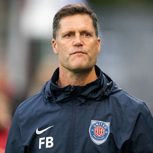 Fredrik Bernhardsson i sportkläder står framför en fotbollsplan. Han bär en jacka med en logo på bröstet och ser ut att vara involverad i en träningssession eller match. I bakgrunden syns suddiga konturer av andra personer och en del av planen.